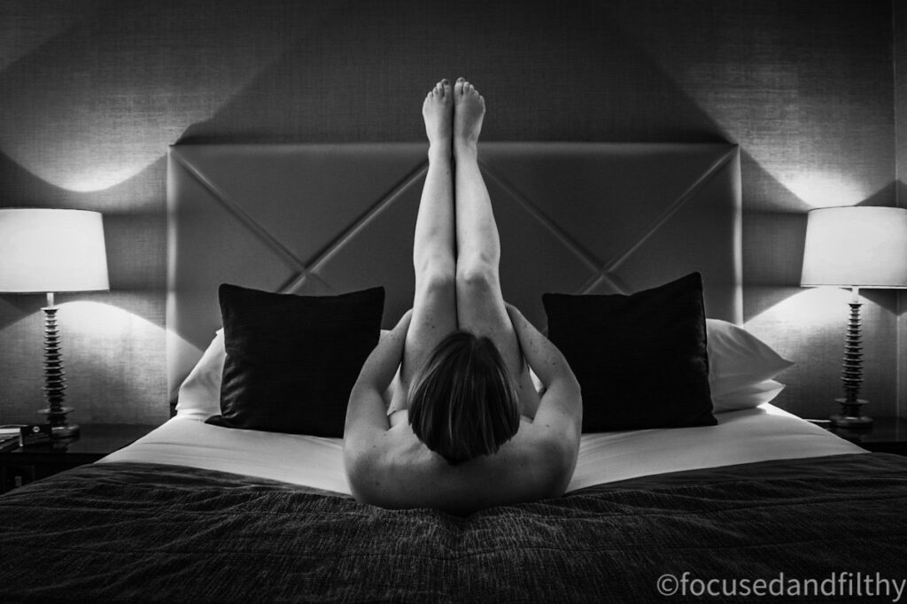 Black and white photograph of naked woman on a bed with her legs straight in the aor dacinf away from you  with cushions and lights either side   