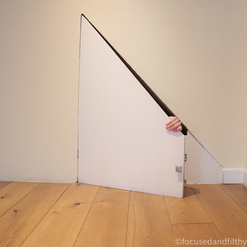 Colour photograph of a small triangular cupboard woth just a hand holding the door shut 