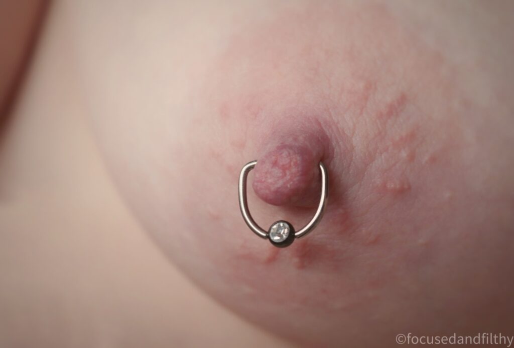 Close up colour photograph of a left naked breast woth a silver D shaped nipple ring in that has a black ball closure with a clear sparkly jewel in  