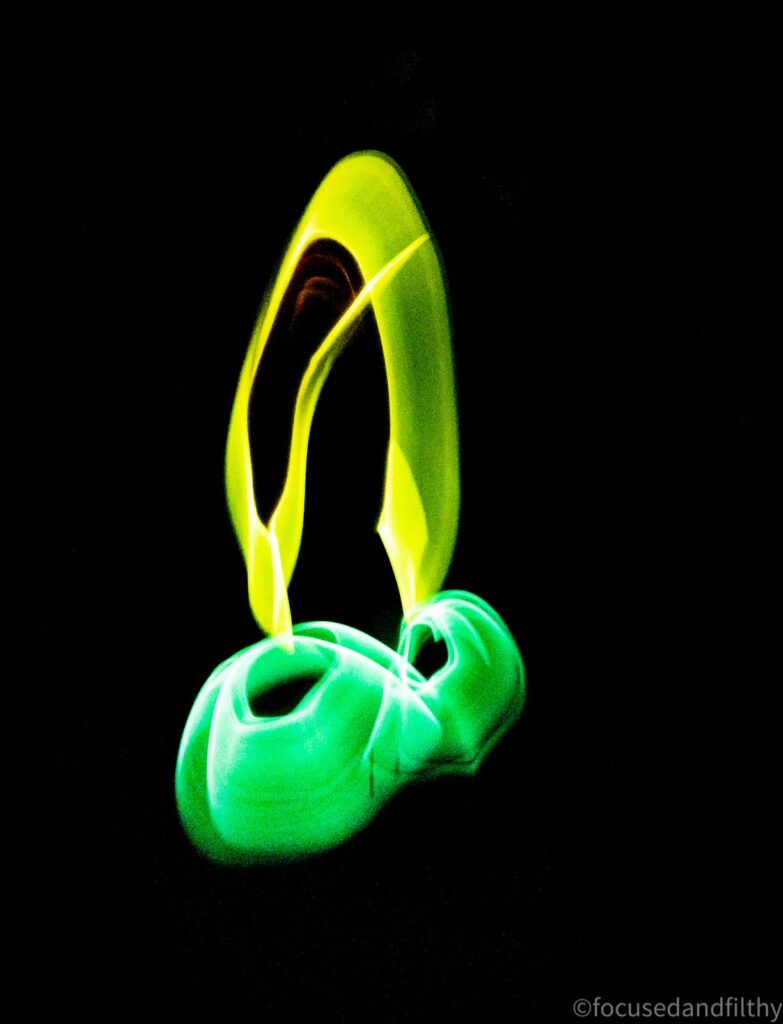 Dark photograph depicting a cock and balls but with yellow and green glow sticks on a long exposure. 