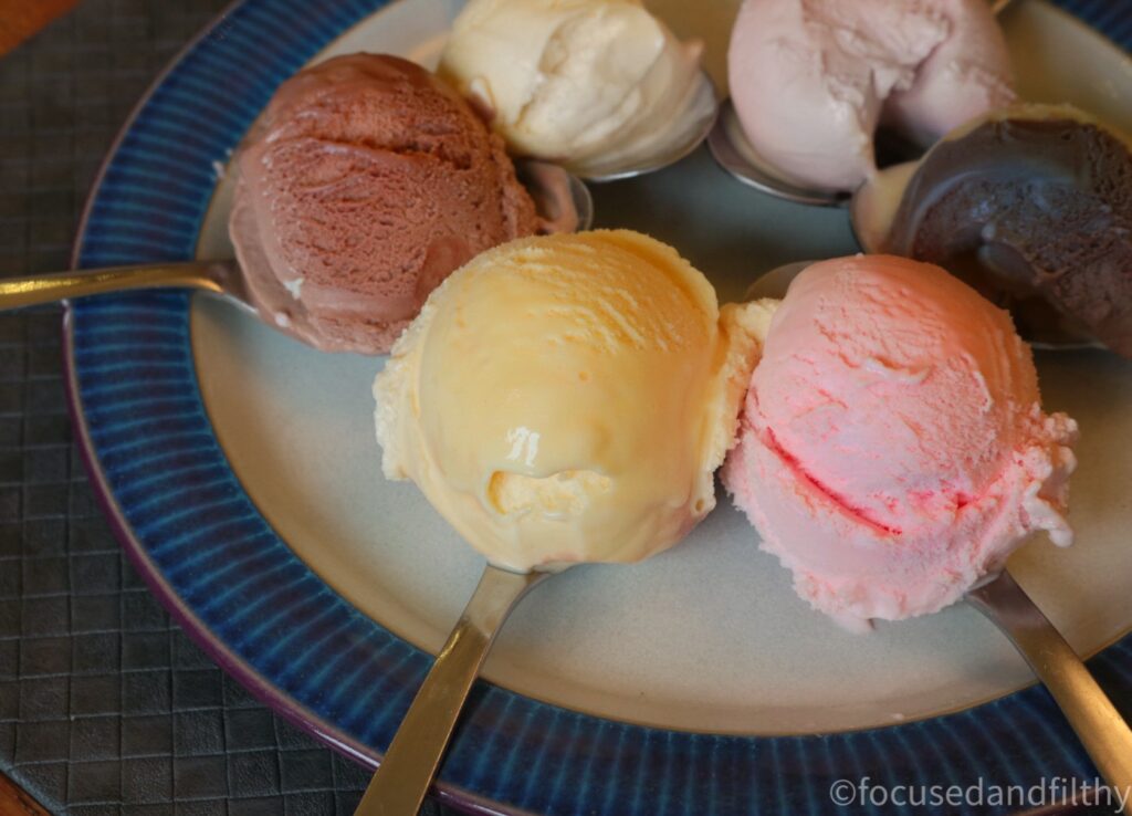 A colour photograph of a plate on a table with six spoons on it. Each spoon has one scoop of different coloured and probably flavour of ice cream on it. The are arranged like a star with the handles pointing outwards. 
