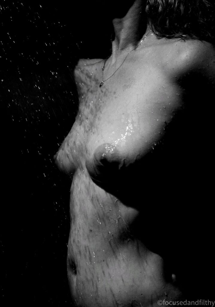 Black and white photograph of the torso of a naked woman in the shower with the water hitting her breasts. 