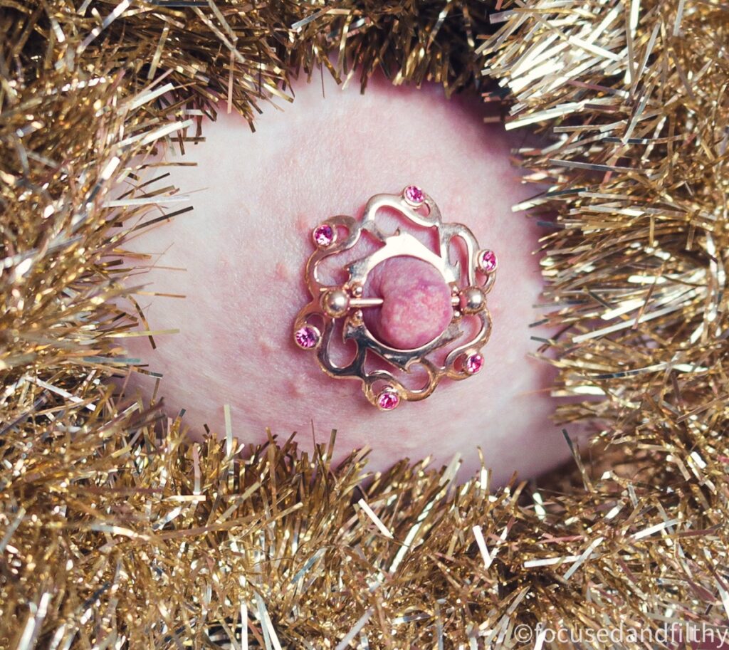 Close up colour photograph of a left nipple surrounded by gold tinsel. The nipple has a large rose gold nipple shield with 6 sparkly pink stones set in it and is held in place with a rose gold nipple bar. 