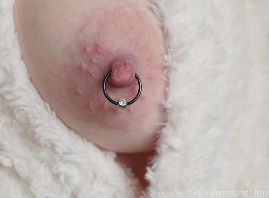 Close up colour photograph of a naked left breast peeking out of a white fluffy dressing gown. The nipple has a large black nipple ring through it with a sparkly ball closure. 