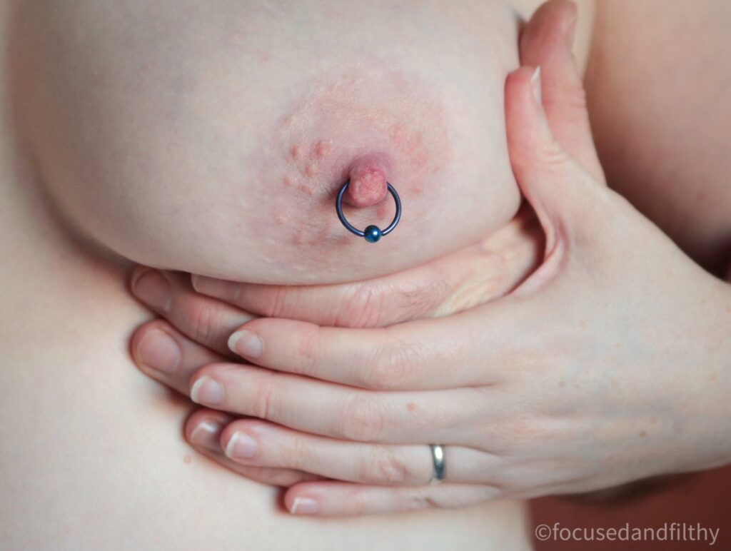 Close up colour photograph of a naked left female breast with two hands underneath. One male and a smaller female hand over the top. The hand is cupping the breast. The nipple has a large dark blue nipple ring in it. 