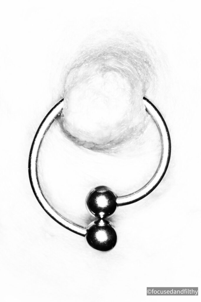 Very close up black and white photograph of a nipple that has a nipple ring in. The ring has two balls that sit directly on top of each other. Almost like a spiral but not quite. 