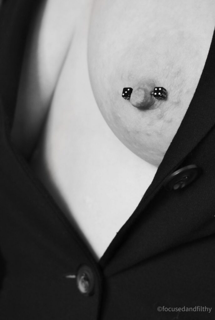 Black and white close up photograph of a female breast in a black jacket with nothing underneath. The left nipple is exposed and shows a nipple bar that has black dice on either end with white spots for numbers on. 