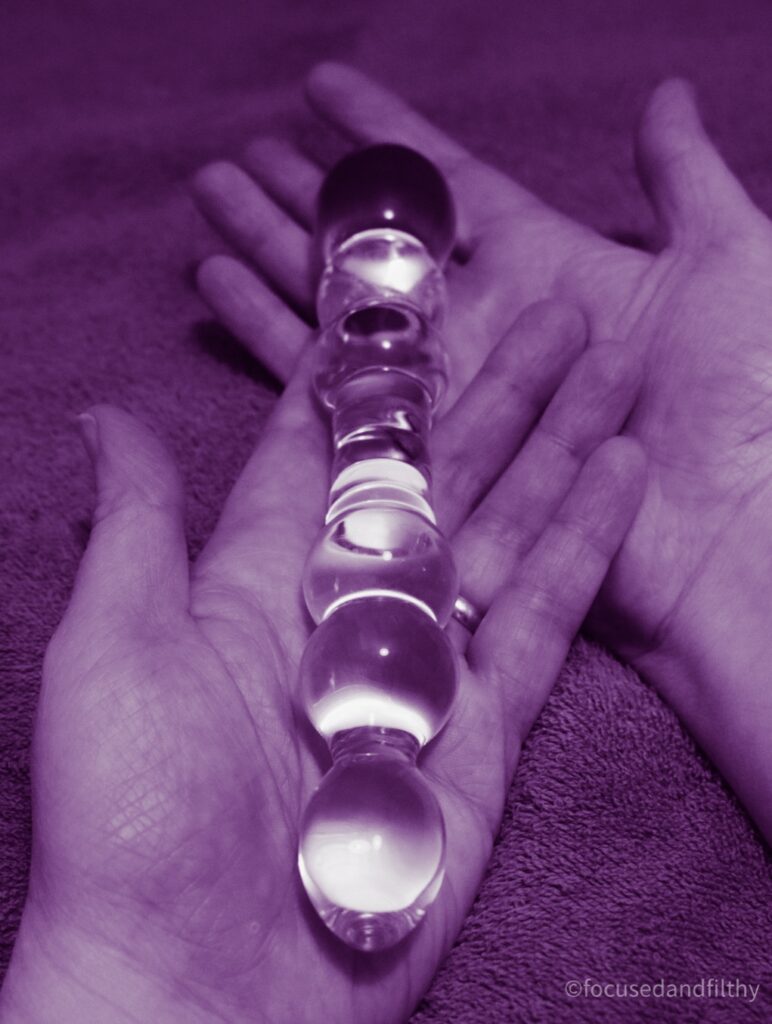 Close up photograph of a glass bulbous dildo sat on two open hands. The photograph is tinted so it all looks purple. 