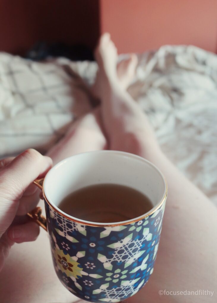Colour photograph of my point of view looking down onto my bed I’m holding a blue patterned mug over my mug and you can see my naked pale legs in front of me. 