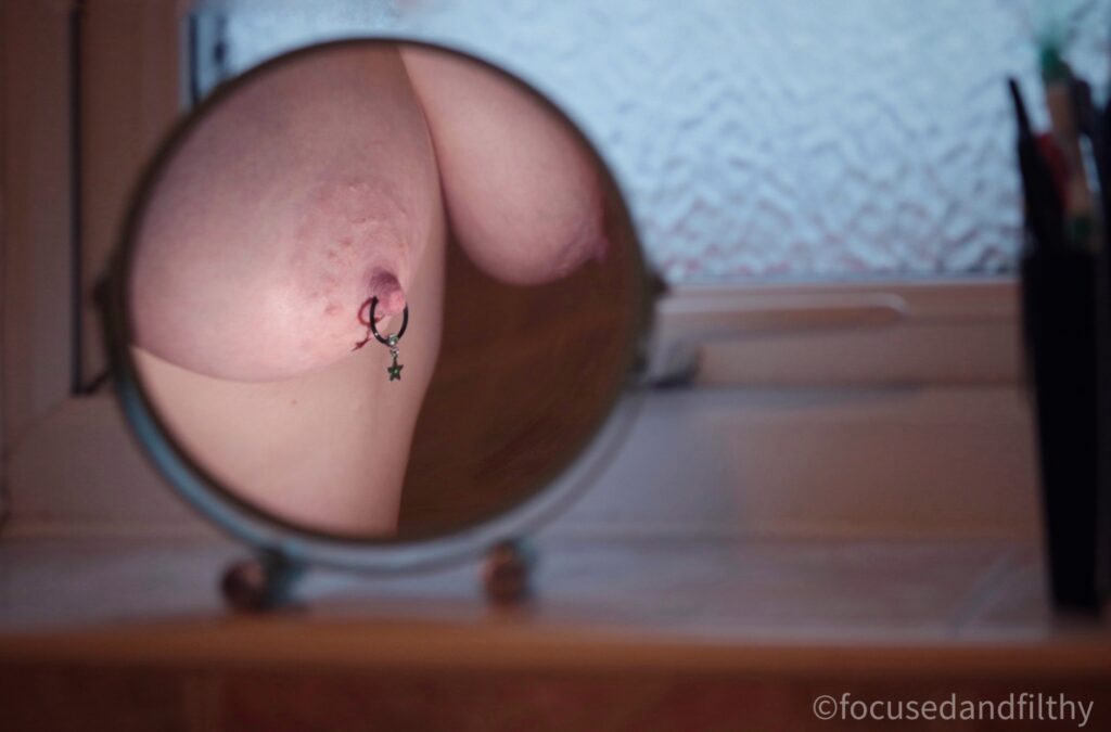 A colour photograph of a bathroom windowsill which has a small round mirror on it. Reflected in the mirror is a bare of women’s naked boobs. The left nipple has a round black nipple ring in with a ball closure and a small star dangling from it. The window is relatively dark but the breasts are well lit. 