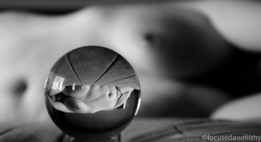 A black and white close up photograph looking through a lens ball showing a naked woman lying on a bed her torso is in focus and upside down in the lens ball but you can see the out of focus body behind the ball. 