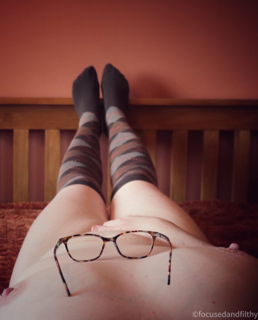 Colour photograph from my eye view. Looking down my naked body on a bed. My tortoiseshell glasses are balanced on my chest. I have one hand over my muff and I’m only wearing long over the knee brown diamond patterned socks. 