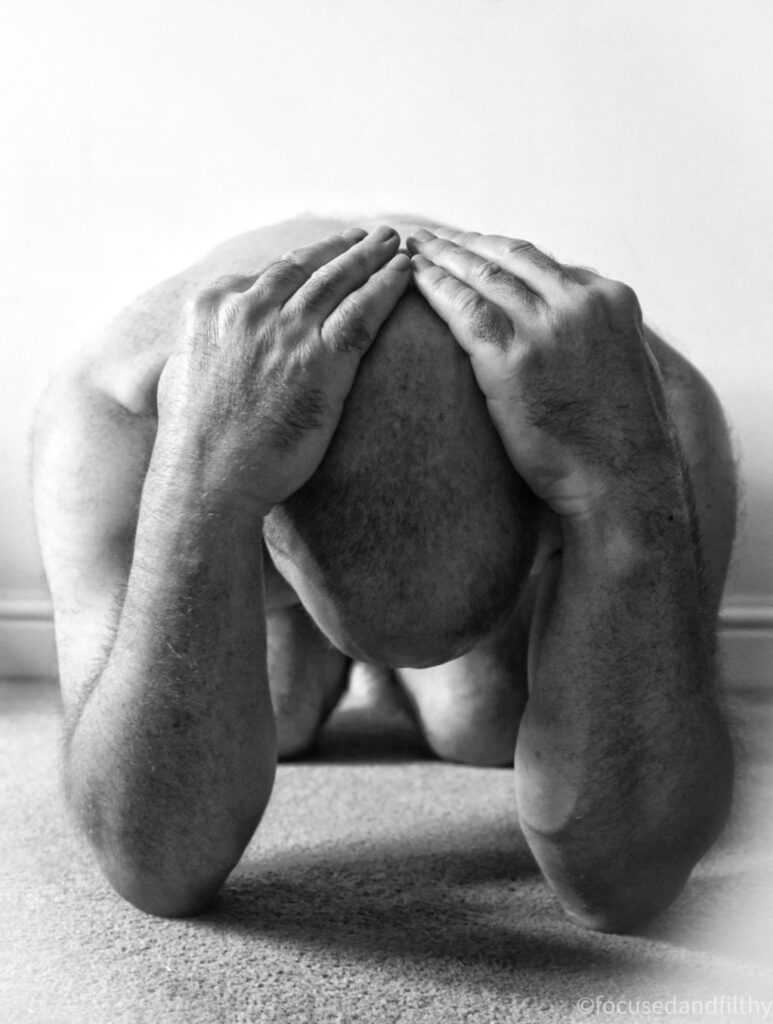 Black and white photograph from a low down image showing a naked man kneeling with his elbows on the ground and his hand in his head. 