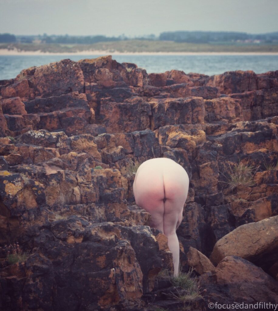 A colour photograph of some interesting rock formations next to the seaside with a naked lady bending over so you can only see her arse and legs. 