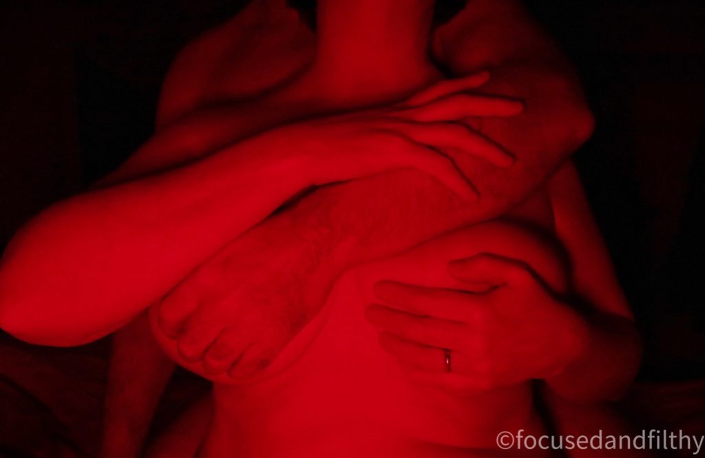 A colour photograph but only taken using a red light showing a naked female torso with a male arm coming over her left shoulder and holding her right breast at a diagonal.  She is holding his arm with her right hand and her left hand is covering her left breast. The image is cropped at the waist and the neck. 