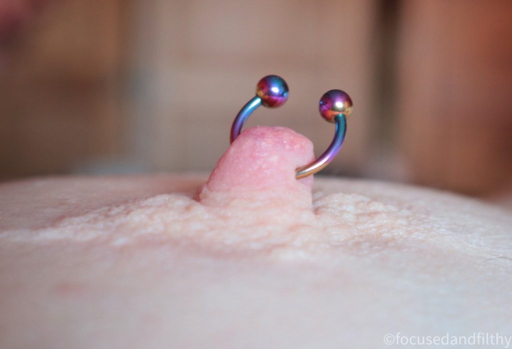 Close up colour photograph of a female nipple with a rainbow colour nipple horseshoe piercing in it.  The nipple is quite erect with the two ends of the horseshoe standing up. 