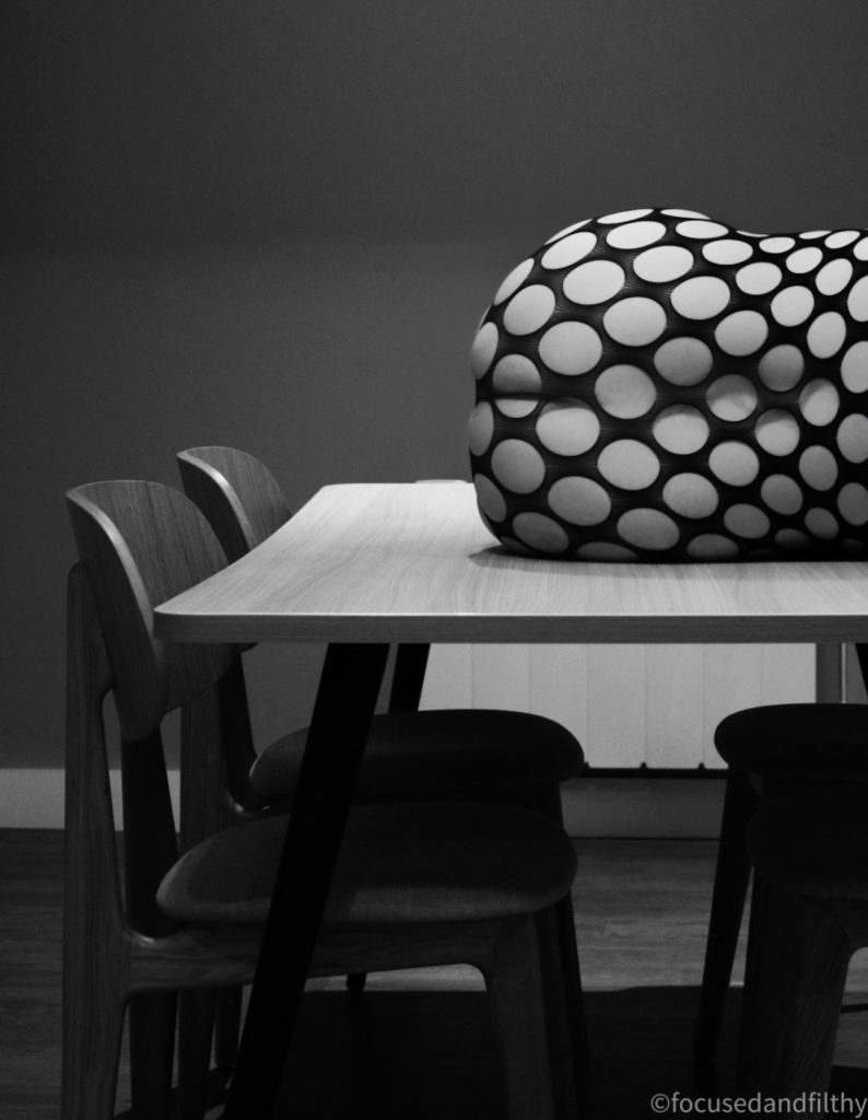 A black and white photograph slightly off centre showing a wooden dining table and two wooden 1950 style chairs. On top of the table is the back and bottom of a naked curved person wearing a fishnet body stocking with large holes in 