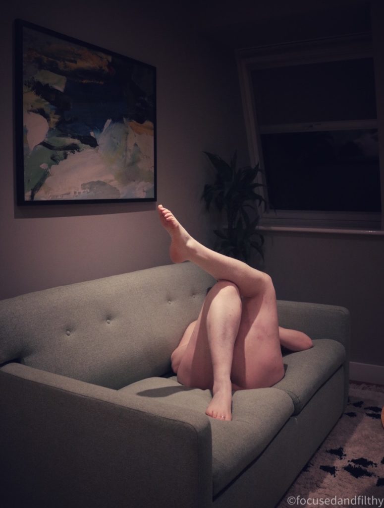 Colour photograph of a naked woman lying on a sofa in a darkened room. She has her legs crossed and one foot is pointing up to the ceiling while the other in on the sofa. You can’t see her face 