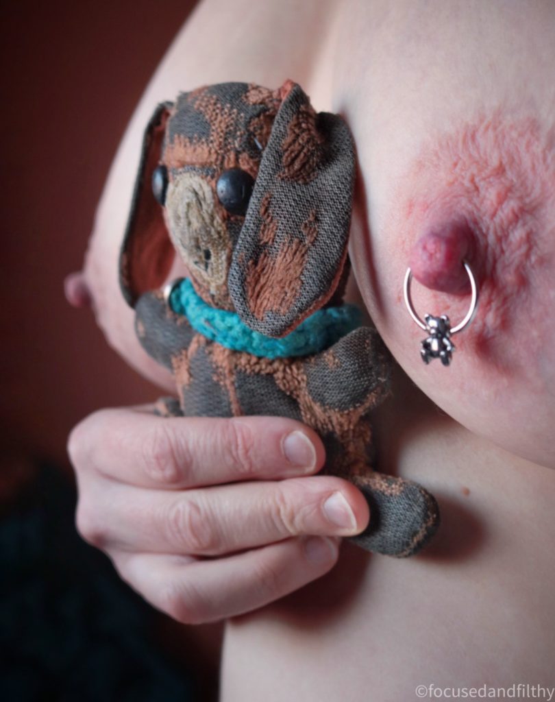 A close up colour photograph of a naked woman’s chest and a small rather worn and tatty looking old brown teddy bear held between the naked breasts. The left nipple has a silver nipple ring in and the closure is a small silver teddy bear shape. 