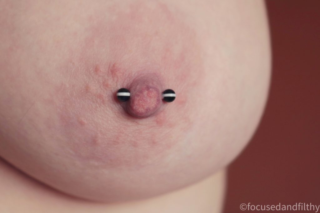 Close up colour photograph of a naked left female breast showing the nipple with a nipple bar in. The balls on the nipple bar are black with one white stripe in the middle.  
