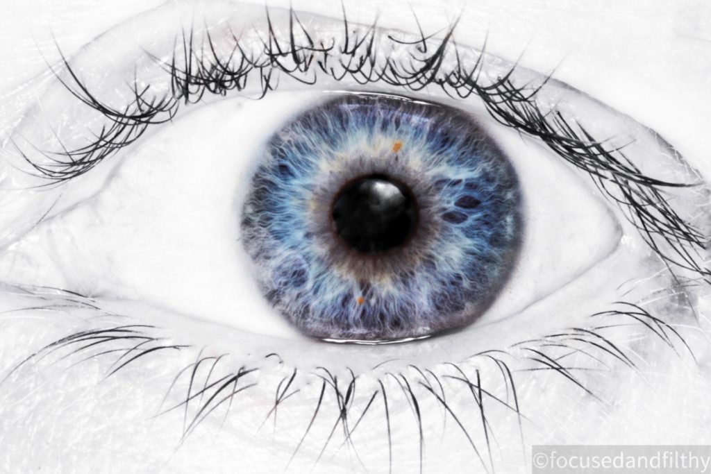 Close up photograph of an eye. The image is mainly black and white but the iris is blue and their are patterns like flames in the iris. The eye lashes are long and dark 