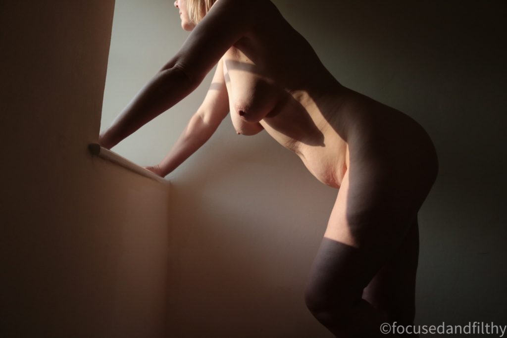 a colour photograph looking up some stairs at a naked woman who is standing with her hands on a windowsill looking out towards a sunny afternoon. The shadow of the windowsill cuts across the top of her thighs making it look like she’s wearing stockings. 