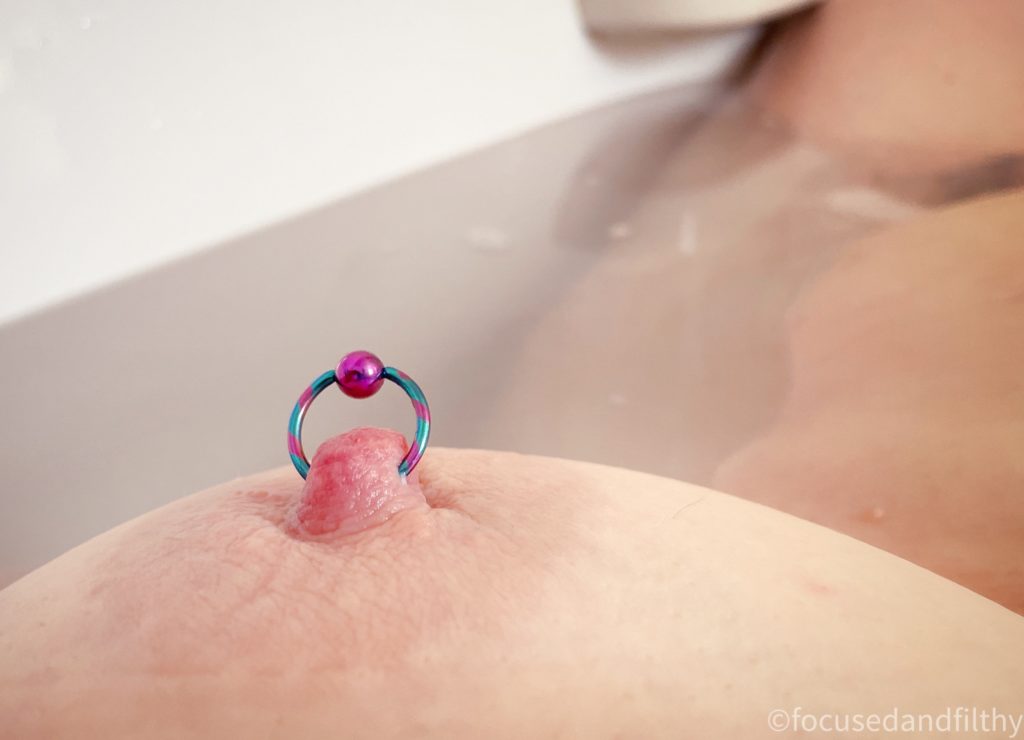 A close up colour photograph of a naked left breast of a woman laying in the bath. The nipple has a turquoise and pink stripped nipple ring in with a large pink ball closure. The water is seen lapping up against her but there is a view of a bit of tummy and thigh in the shot too. 