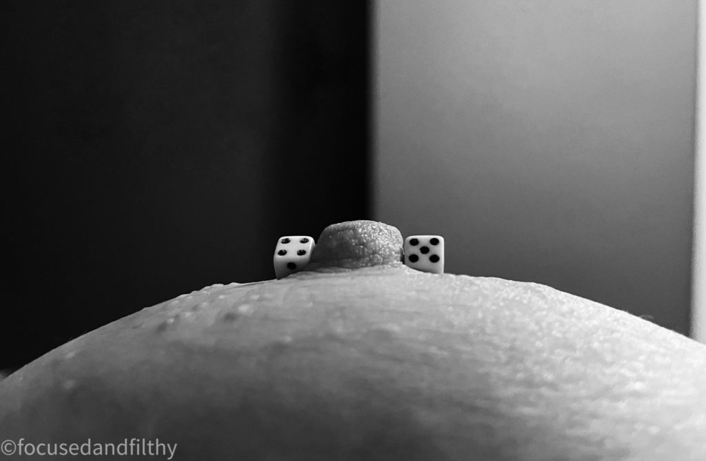 A close up black and white photograph of a left female breast seen almost from her view point showing a nipple bar with white dice on either end. One side has a number 4 and the other a number 5 the back ground is plain but there is a wall line which is right in the midline of the nipple 