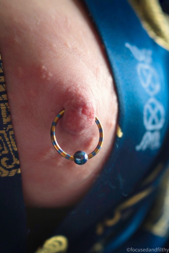 Close up colour photograph of a left nipple with a large blue and gold striped nipple ring in it. The ball closure is blue. The colours match perfectly to the silk dressing gown around the nipple which is blue with gold Chinese patterns on it. 