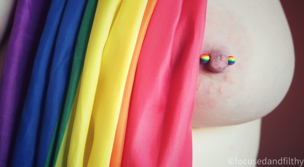 A close up colour photograph of a naked left breast with a rainbow nipple bar in and draped next to it is a pride flag showing all the rainbow colours. 