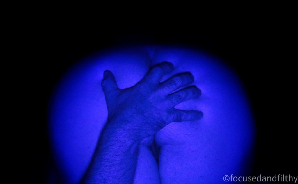 Close up colour photograph of a naked bottom with a male hand grabbing one of the cheeks. The whole image has been taken under a blue light so everything is blue and the background is black. 