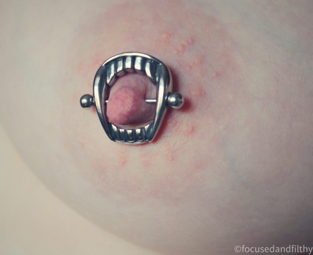 A close up colour photograph of a naked female breast. The left nipple has a nipple bar through it with a silver set of teeth around it like it’s being bitten.  It’s quite large. 