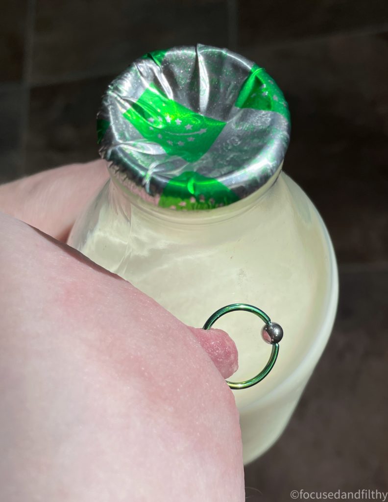 A close up photograph looking down at a bottle of milk being held close to a naked left breast. The milk bottle has a green and silver foil lid and the naked breast has a nipple piercing. The ring is the same colour green as the bottle top with a silver ball closure 