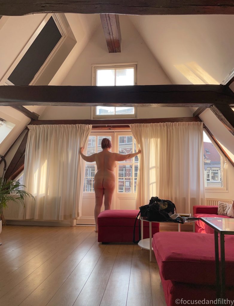 A colour photograph of the inside of a tall Dutch townhouse attic with very steep pitched roof, and dark wooden beams going across. The walls are white, the floor is wood and the sofa is red at the ems of the room is a massive window like double doors with a thin sheet curtain across it and light streaming in on the naked woman who is stood there holding the curtains apart and looking out. You can just see other tall buildings across the road. 