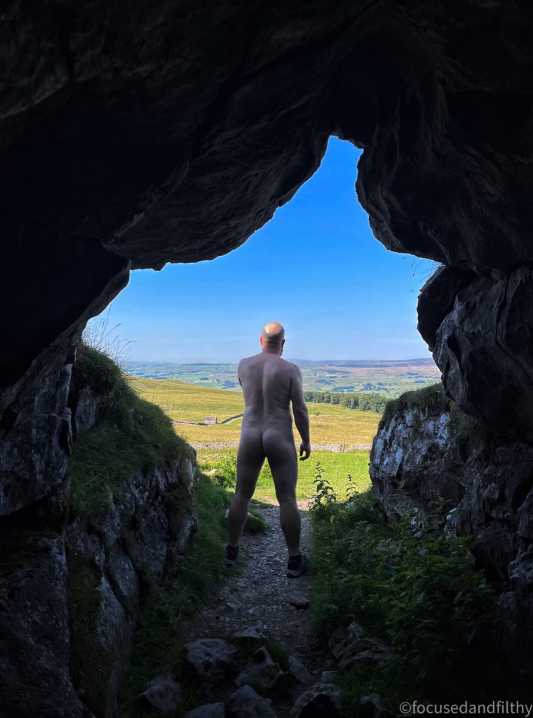 Colour photograph looking out of a dark cave into bright sunny Yorkshire countryside. Stood at the mouth of the cave is a naked man facing out into the sunshine. With strong legs and a muscle-y back.  