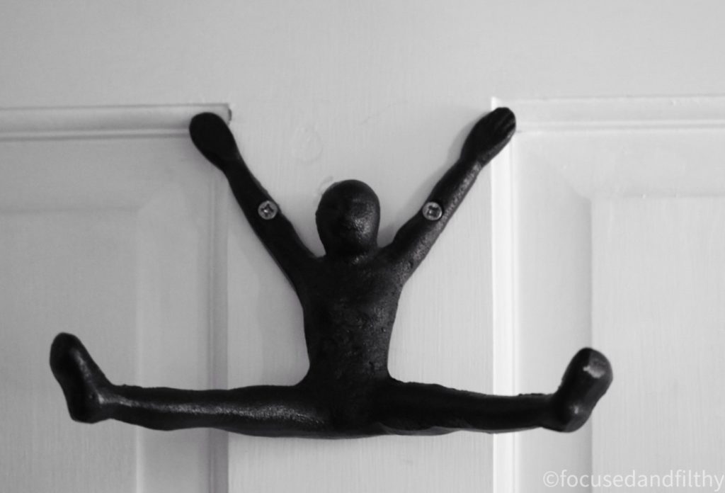 A close up black and white photo of a hook on the back of a bathroom door. But the hook is black metal in the shape of a human with the arms up screwed to the door and the legs out front open wide. 