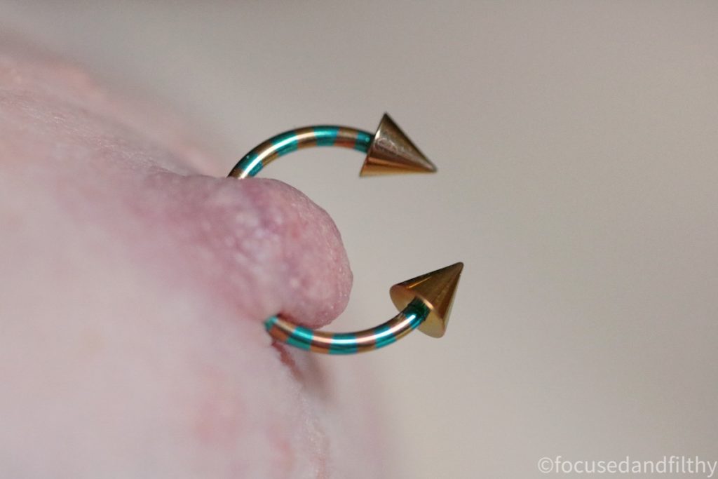 A very close up photograph of my left nipple with a horseshoe shaped nipple bar through that is gold and turquoise striped and on the ends are gold pointy cones. 