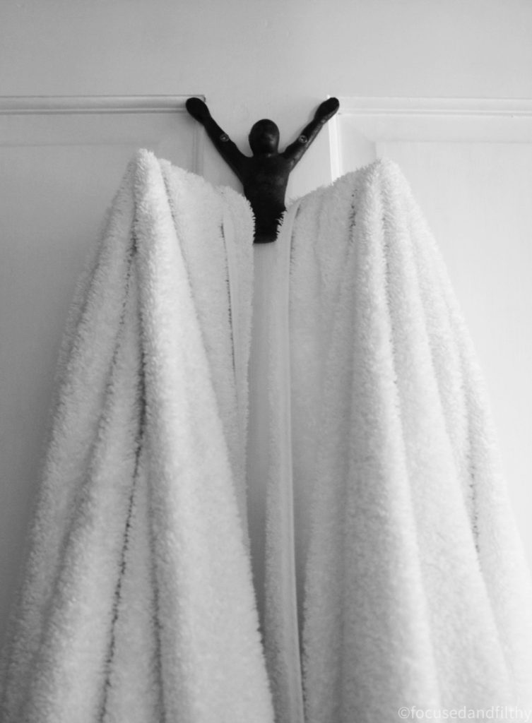 Black and white photograph of the above described bathroom hook (which is the shape of a human attached to the door by their arms and their legs sticking out in front open wide) but with two white towels hanging from the legs. 