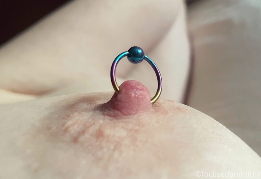 Close up colour photograph of a left nipple with a colour nipple ring in that goes from gold to pink to blue with a blue ball closure on it. 