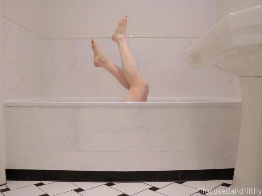 A colour photograph of a rather white bathroom, showing a white bath, white walls, a white sink and black and white checked flooring. Sticking up out of the bath and two naked female legs pointing their toes upwards 