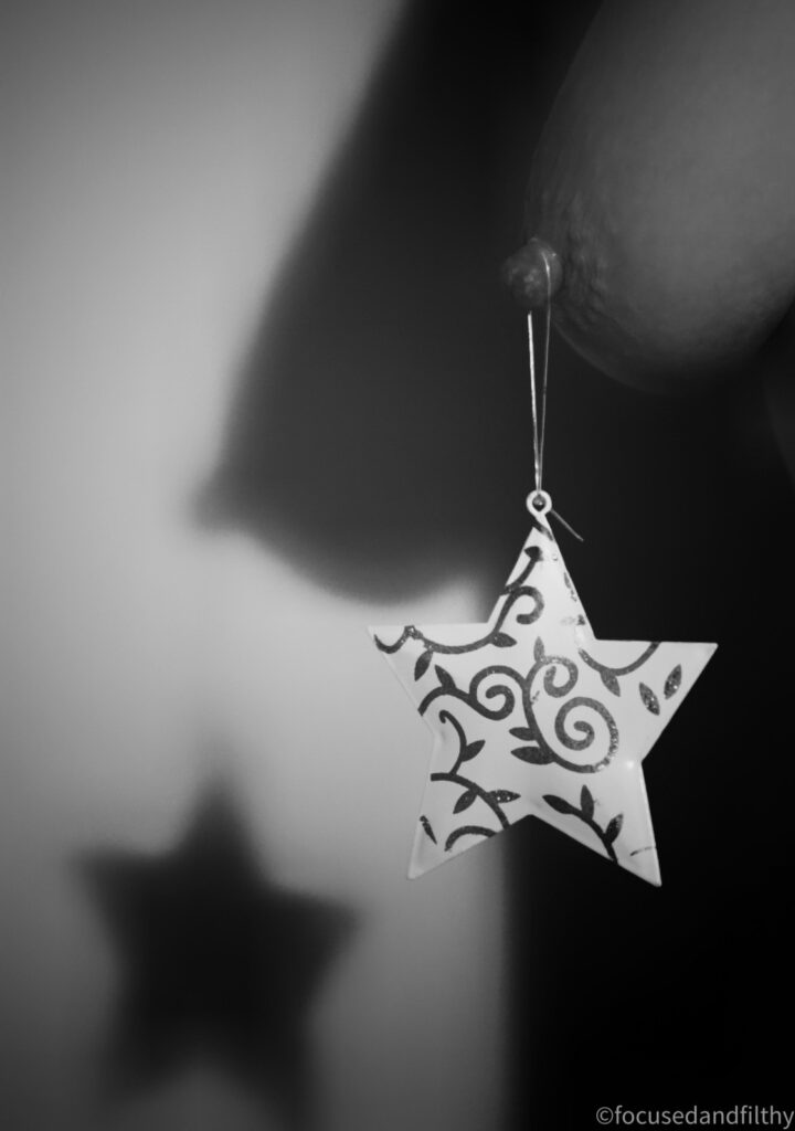 Black and white photograph of a naked breast with a star Christmas decoration hanging off the nipple the star is white with shiny swirly patterns painted on it. The image is taken near a wall and the shadow shape of the breast and star is seen behind her. 