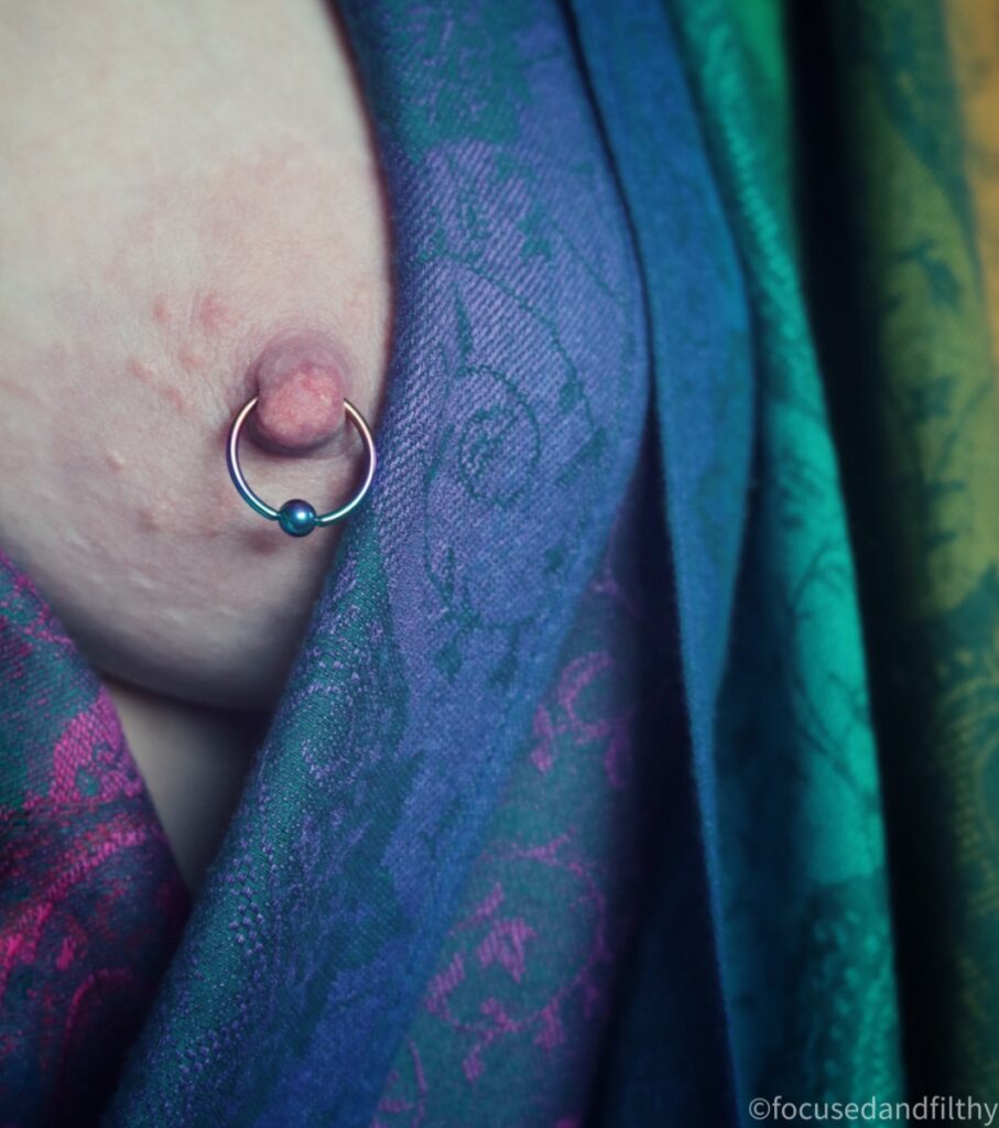 A close up colour photograph of my naked left breast with a rainbow coloured pashmina scarf wrapped next to it.  The nipple has a multicoloured nipple ring in that reflects the colours of the scarf of blue gold and pink and green 