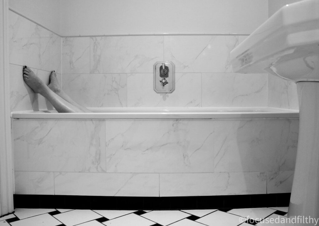 Black and white photograph of a bath.  It’s large with just two feel sticking up at the end against the wall. The tiles are slightly marbled effect but generally all white with a black and white floor  