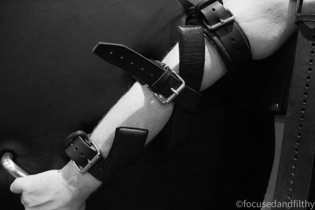 Black and white close up photograph of an arm with several buckets attached around it holding to a padded backing. The hand is grasping a metal bar. 