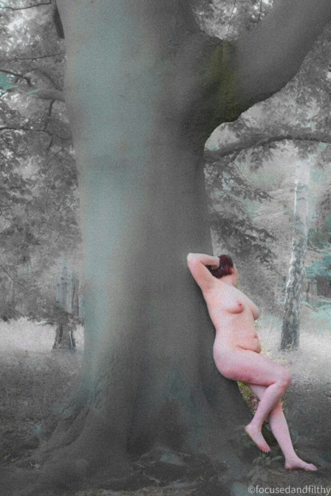 A coloured photograph with a soft dreamy edit feel of a naked woman leaning up against a large beech tree. Showing just her side profile and tilting her head away. 