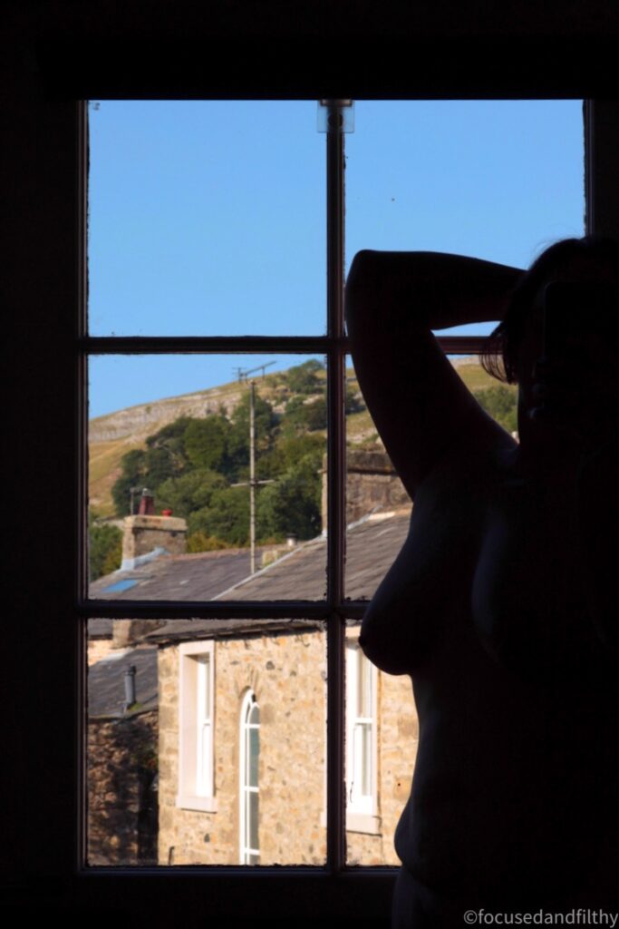 A colour photograph of a silhouetted naked female torso against a windowed door outside the window is a beautiful blue sky and warm honey coloured stone buildings from the dales 