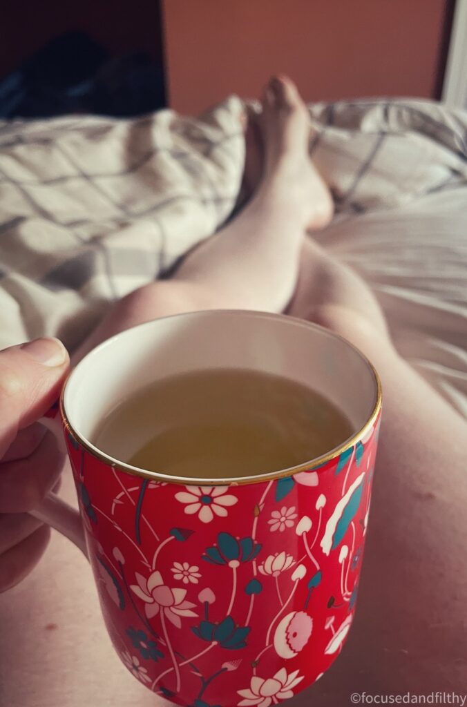 A colour photograph from my perspective looking down and my hand holding a cup of herbal tea in a beautiful red mug and my naked legs stretched out in front of me on the bed 