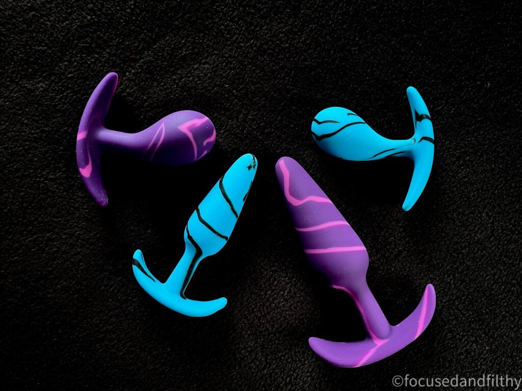 A colour photo of four butt plugs arranged in a semi circle. There are two that have an asymmetrical bump for prostate pressure and two that are smoother and more cone shaped all with bar ends. There are two sizes in both. The smaller ones and blue and the bigger sized ones are purple. They are photographed from above. 