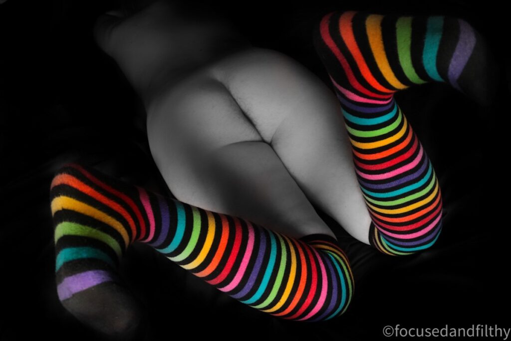 A colour pop image that is black and white apart from the socks. It’s an image of a naked woman from the waist down lying stomach down on the bed with the knees bent and feet flexed. The socks are rainbow coloured and striped. 
