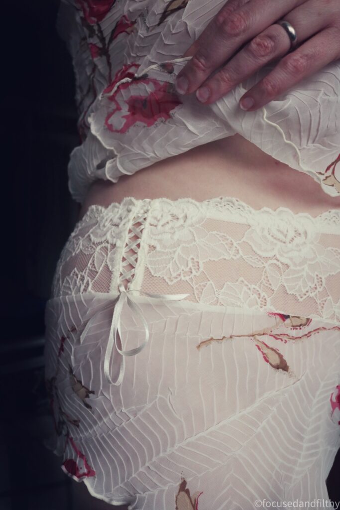 A colour close up photograph of the side of my body wearing a camisole set which is a pale cream with red roses on. The knickers have a thick lace waist bad and the side has a ribbon threaded detail which I’m showing to the camera. 